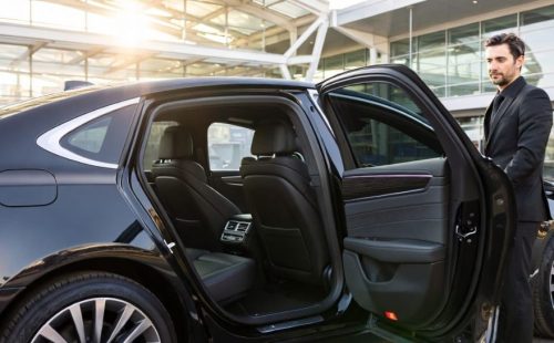 Black executive sedan with chauffeur opening rear passenger door outside a corporate building.