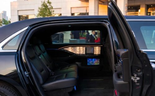 Black stretch limousine with open rear passenger door, showing premium leather interior, ambient lighting, and digital displays.